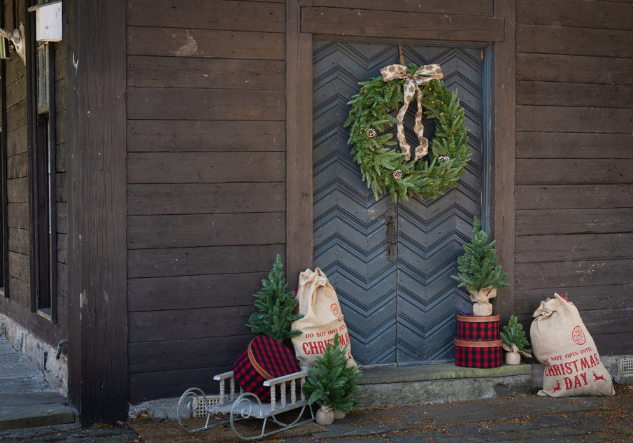 A large Christmas wreath on the door of a wooden cabin with small christmas trees on the veranda and sleighs with christmas bags on them 