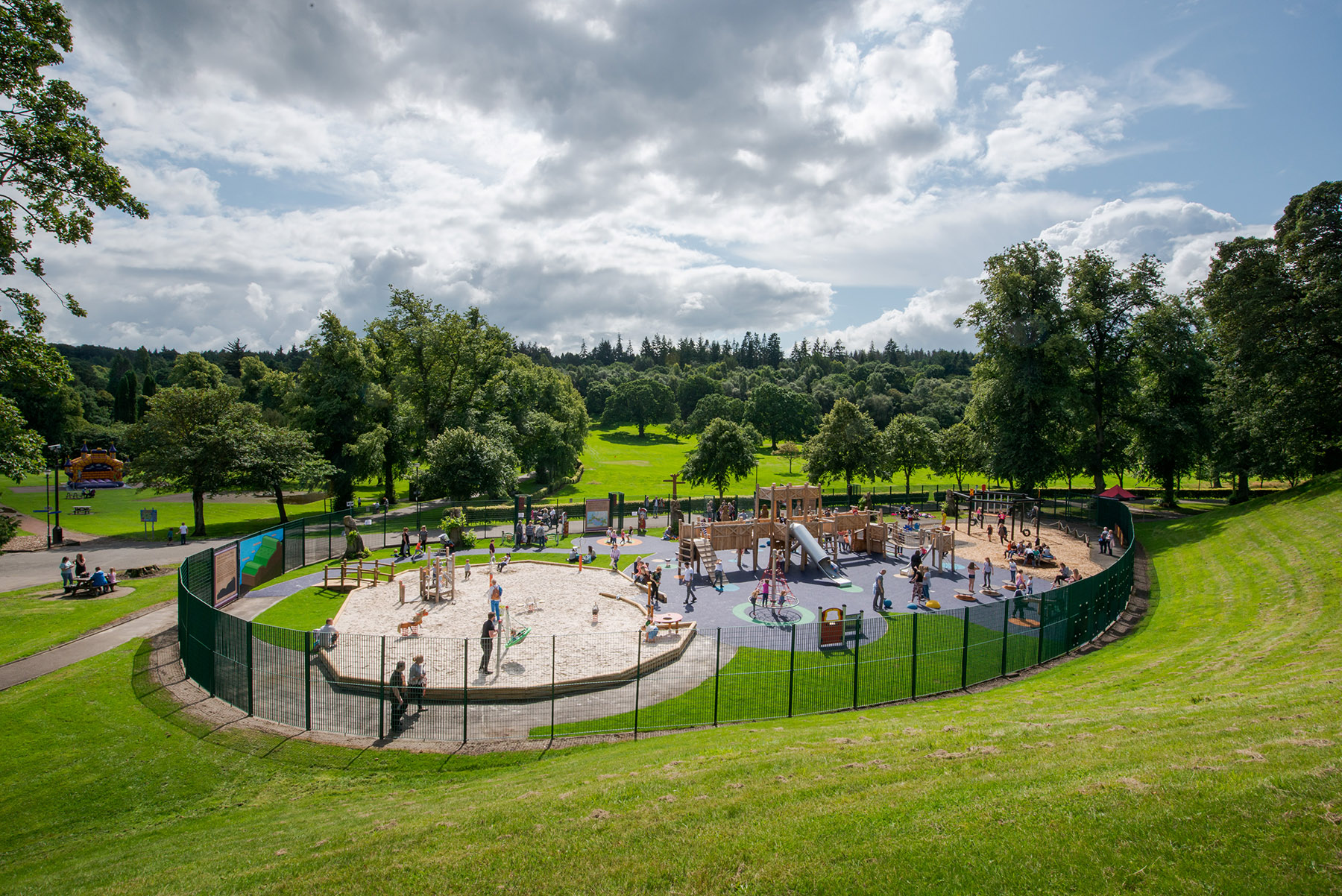 A playground full of children on a sunny day. The playground is surround by a fence and appears to be situated in the middle of a much large park filled with trees.