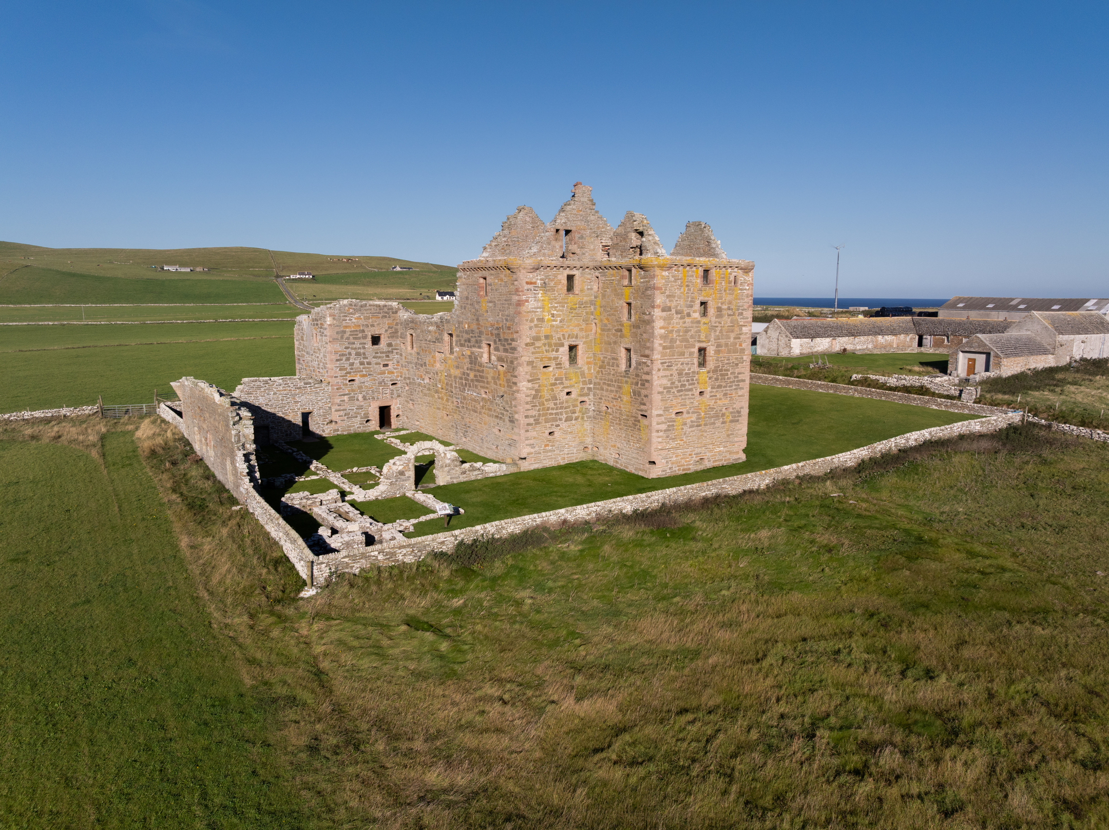 Wide landscape view of Noltland Castle set on open grassland near the coast, with surrounding farmland and scattered buildings.