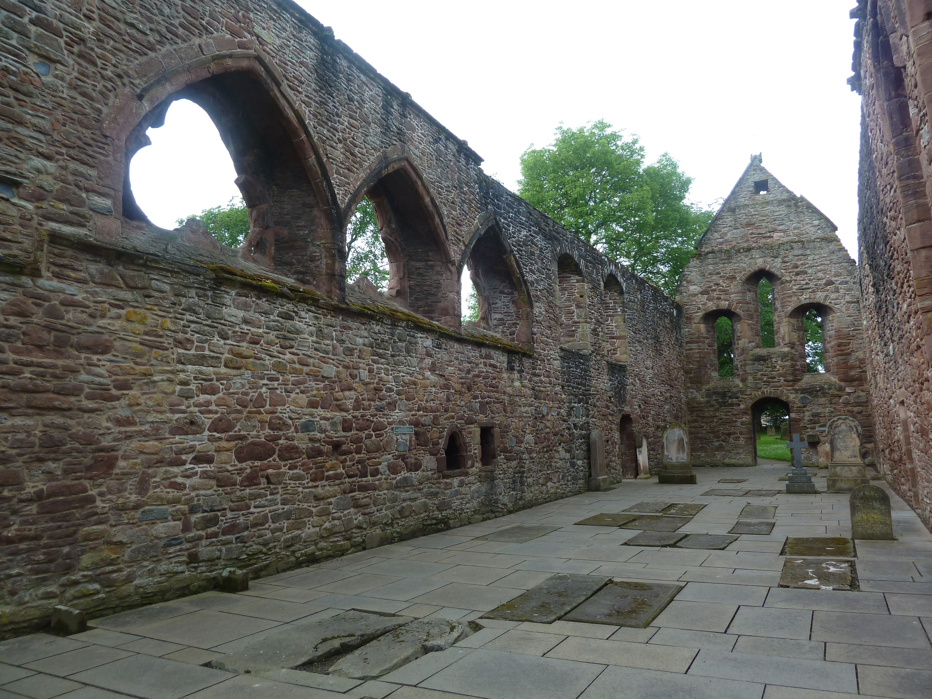 Interior view of the roofless stone church at Beauly Priory, showing long red‑brown sandstone walls lined with tall pointed arch openings. The floor is paved with large rectangular slabs, some of which appear to be grave covers. At the far end, a gabled stone wall contains several narrow arched openings. Trees are visible through the window spaces, and the scene is lit by natural daylight.