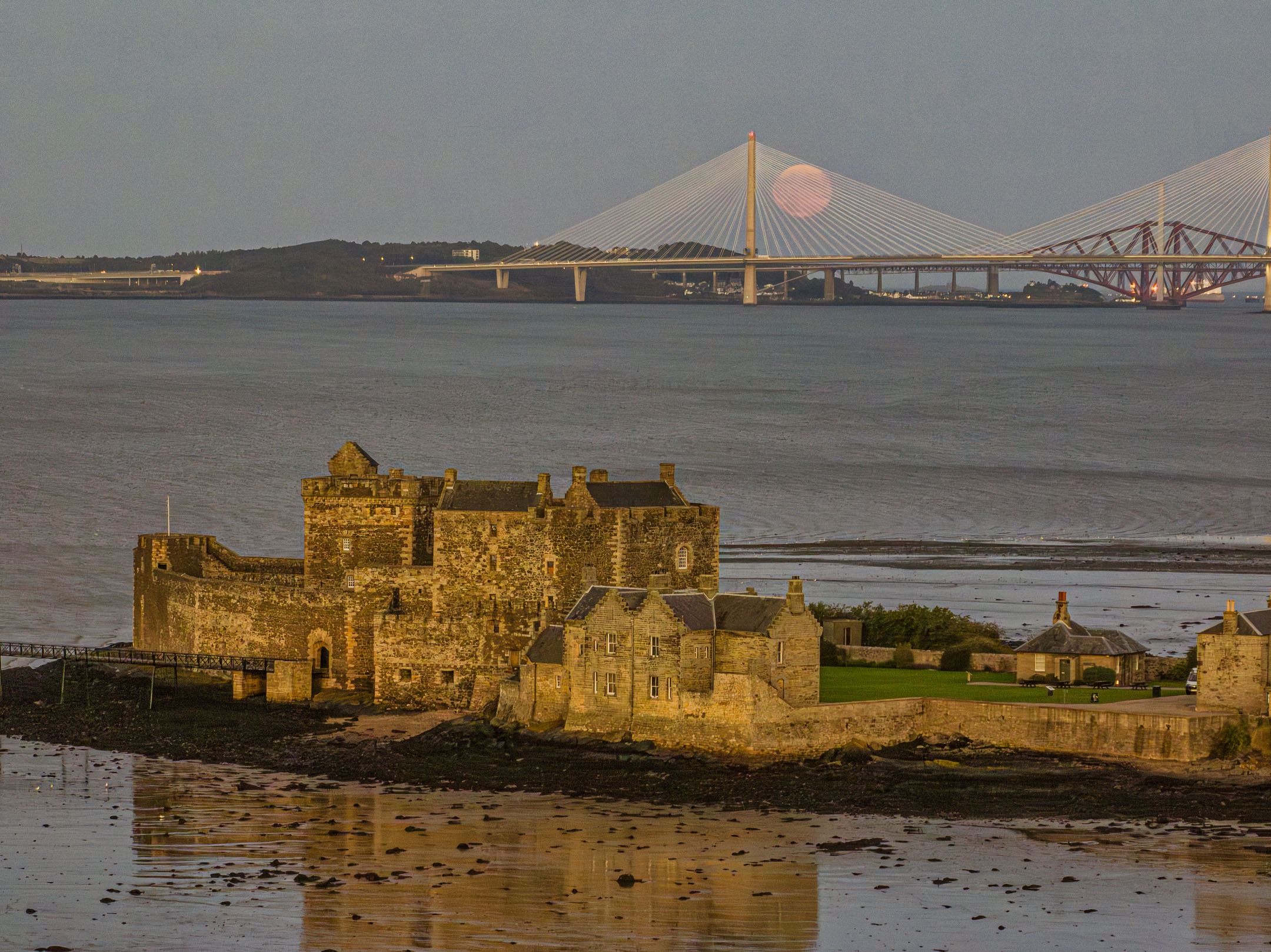 Stone castle on coast with bridges and pink moon in evening sky.