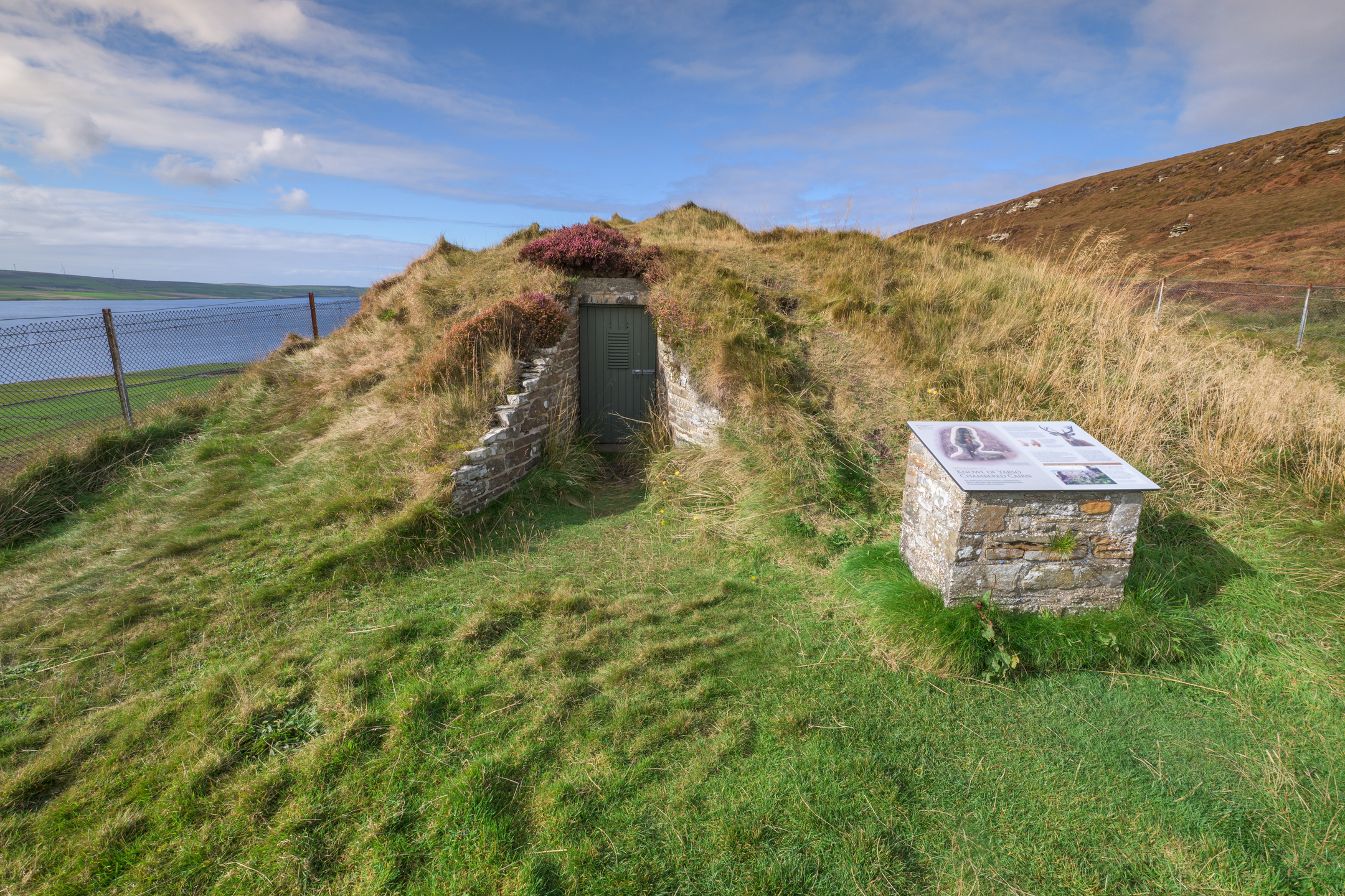 A doorway at the end of a grass-covered mound which gives access to the interior of a chambered cairn. Beside the entrance there is an interpretation panel offering information about the site.