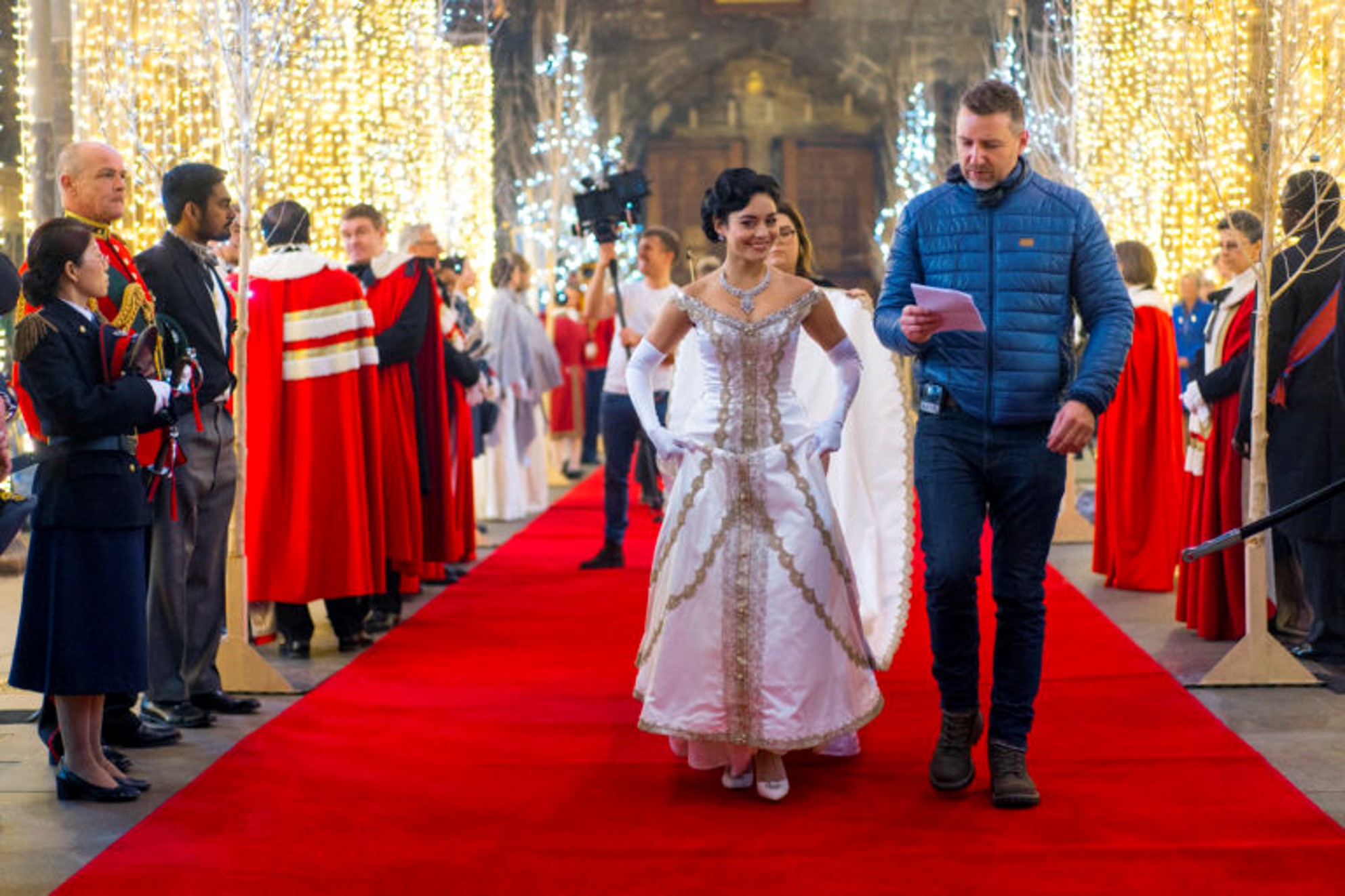 A woman in an elaborate white period costume walks down a red carpet inside a grand hall, guided by a man holding a script, with cast and crew visible on either side.