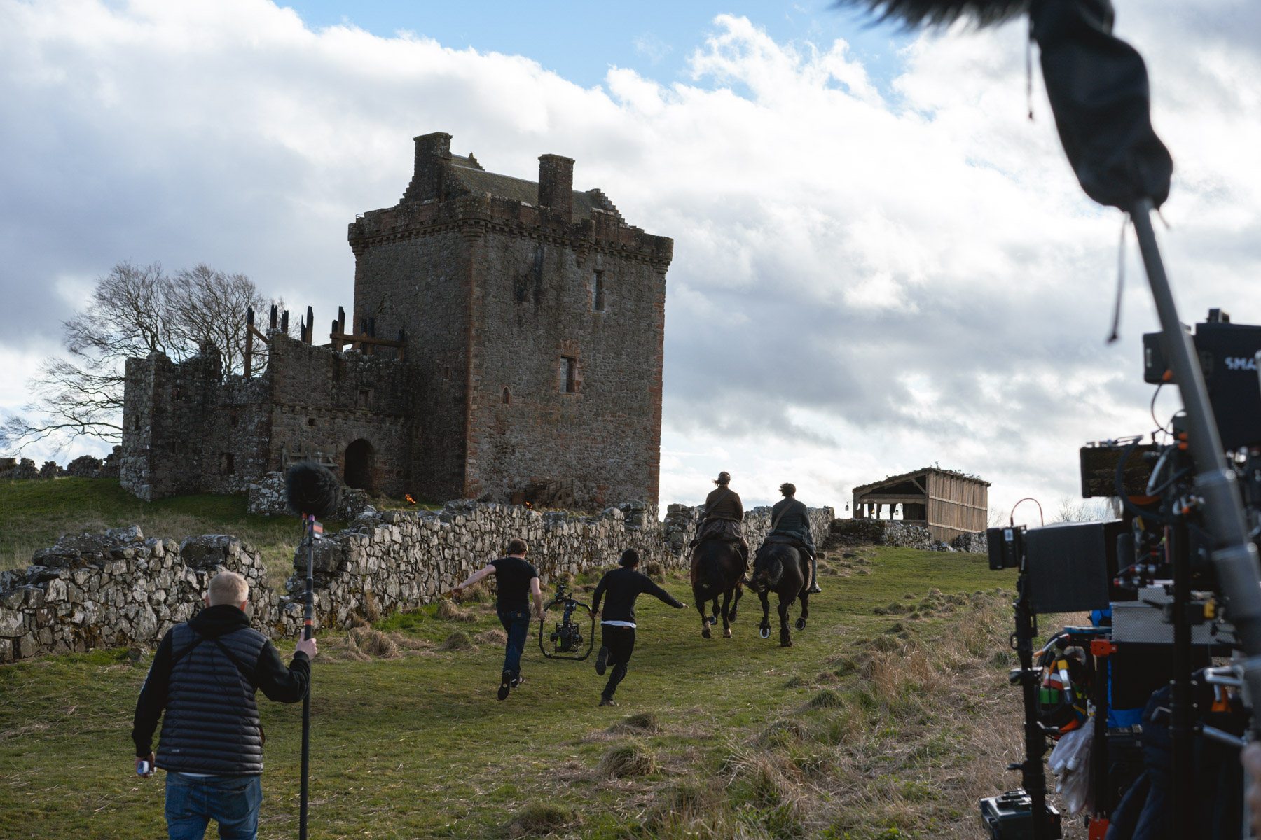 Camera operators follow two actors on horseback as they ride towards a stone tower house.