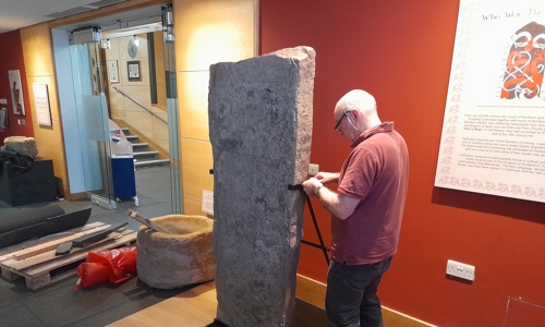 A man fits clamps around an upright Pictish stone, taller than him, to display at the museum.
