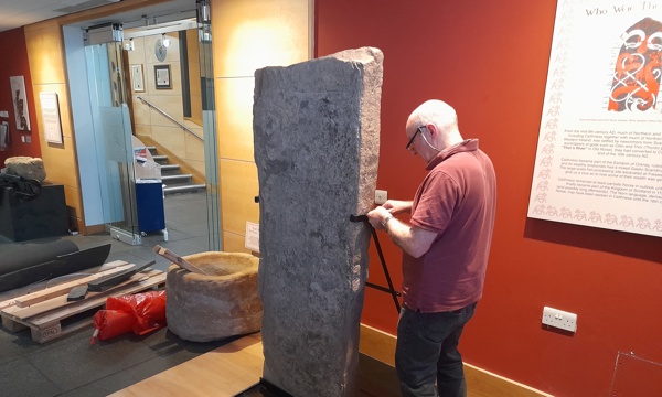 A man fits clamps around an upright Pictish stone, taller than him, to display at the museum.