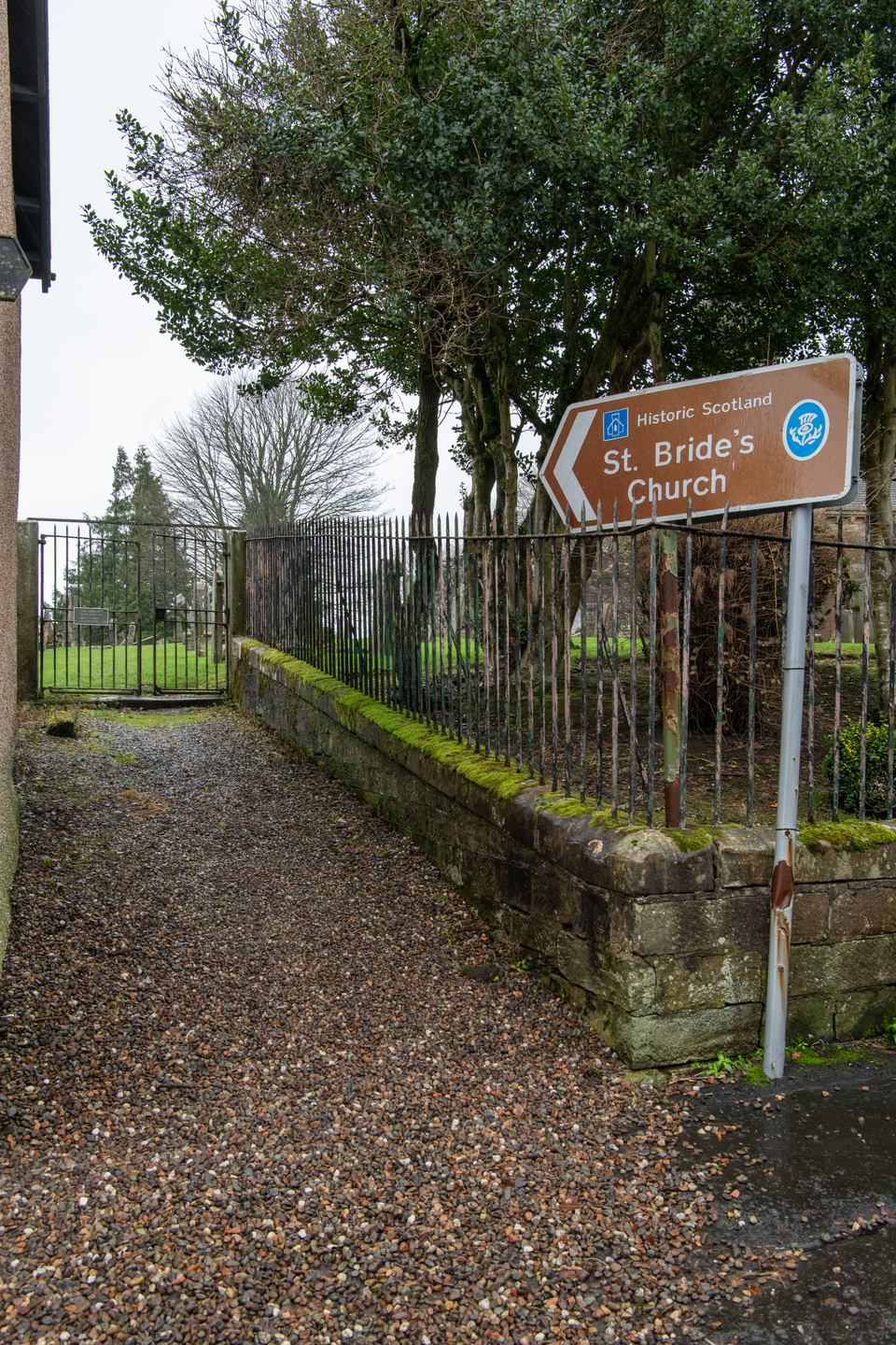 A pathway leading to a metal gate, with a brown Historic Scotland sign reading “St. Bride’s Church” and an arrow pointing ahead. The area is surrounded by trees and stone walls.