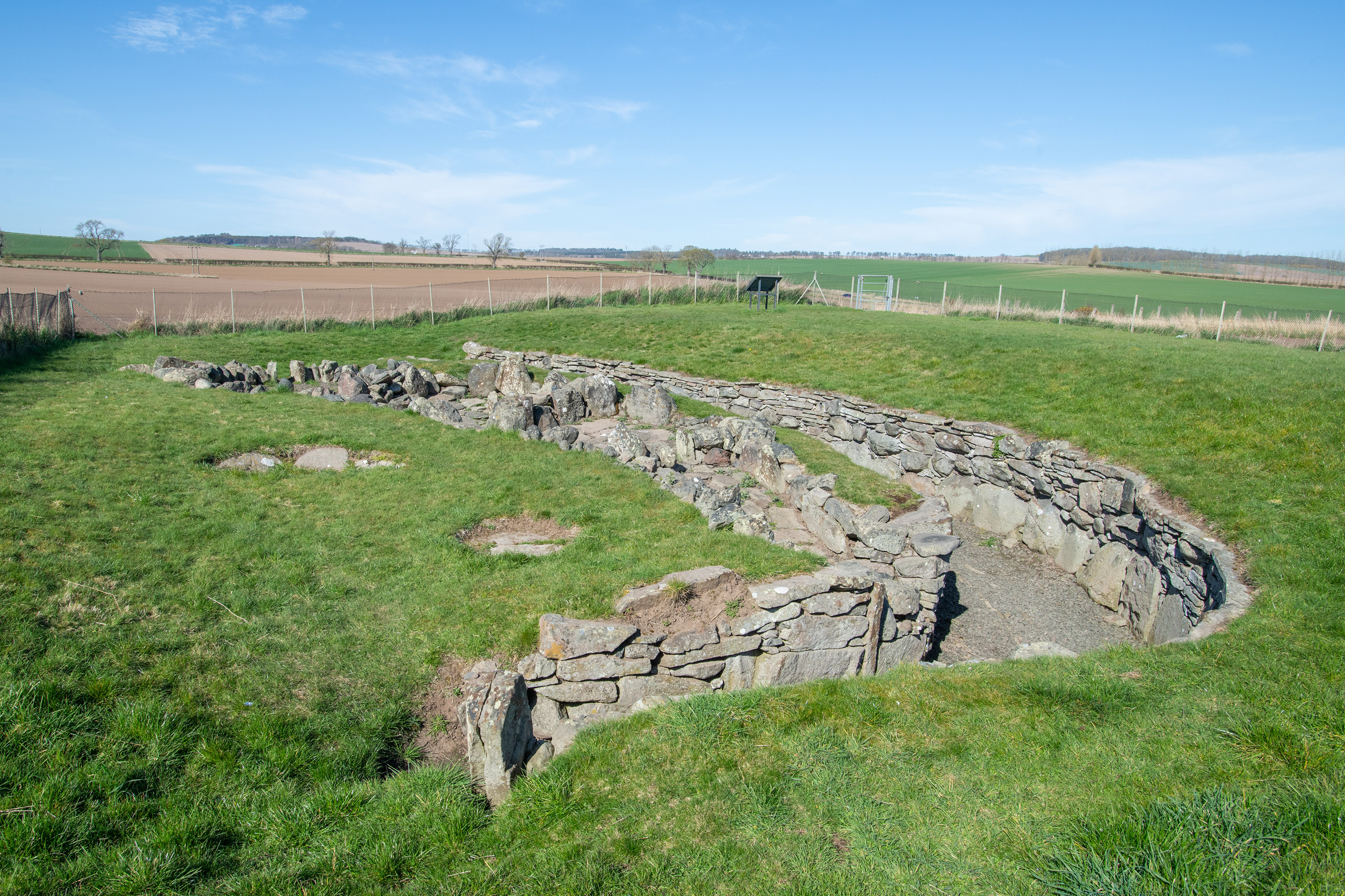 Ground-level view of the curved stone remains of Ardestie Earth House set into a grassy enclosure. The surviving dry‑stone walls form an elongated, winding shape, with sections of the passage dipping below ground level. Beyond the site, wide farmland and gently rolling green hills stretch into the distance beneath a bright blue sky.