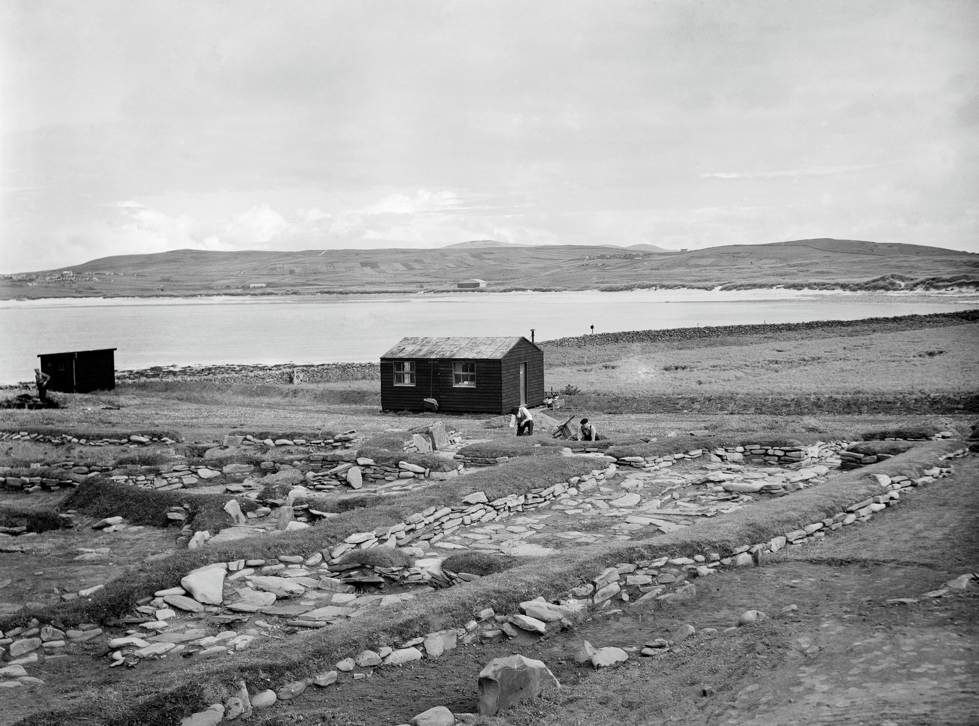 An excavation at Jarlshof, Shetlands. There is a small building in the middle of the photo, and two people in the middle working on the ground. You can see stones on the ground marking a settlement. There is a body of water in the background. 
