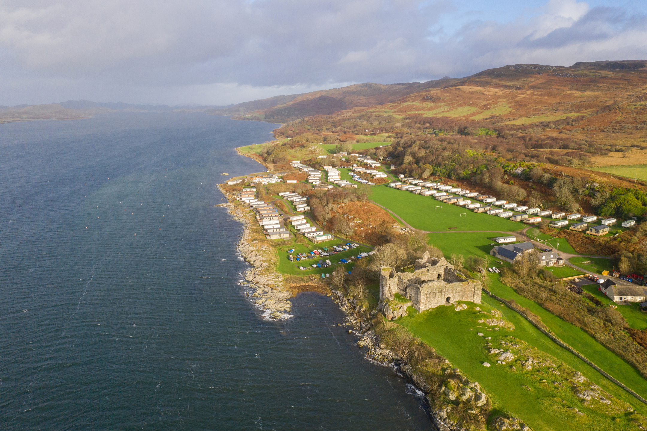 View of water on the left and land on the right, with castle close to shoreline and area with vehicles parked