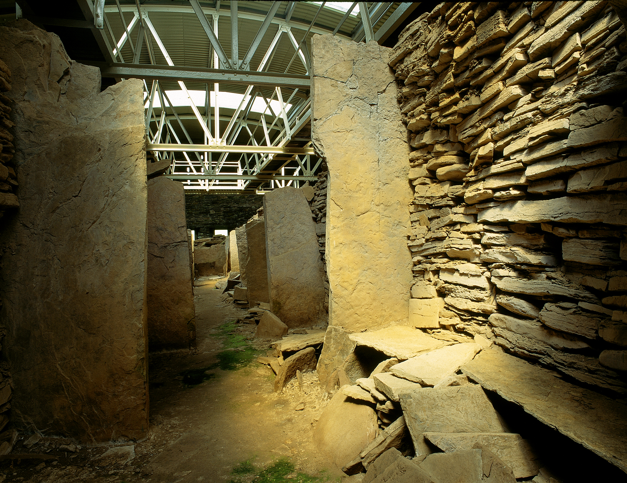A ground‑level interior view of the Midhowe Chambered Cairn, showing tall upright stone slabs forming chamber divisions along a central passage. Stacked stone walls line the space, illuminated by light filtering through the protective modern roof and metal support beams overhead.