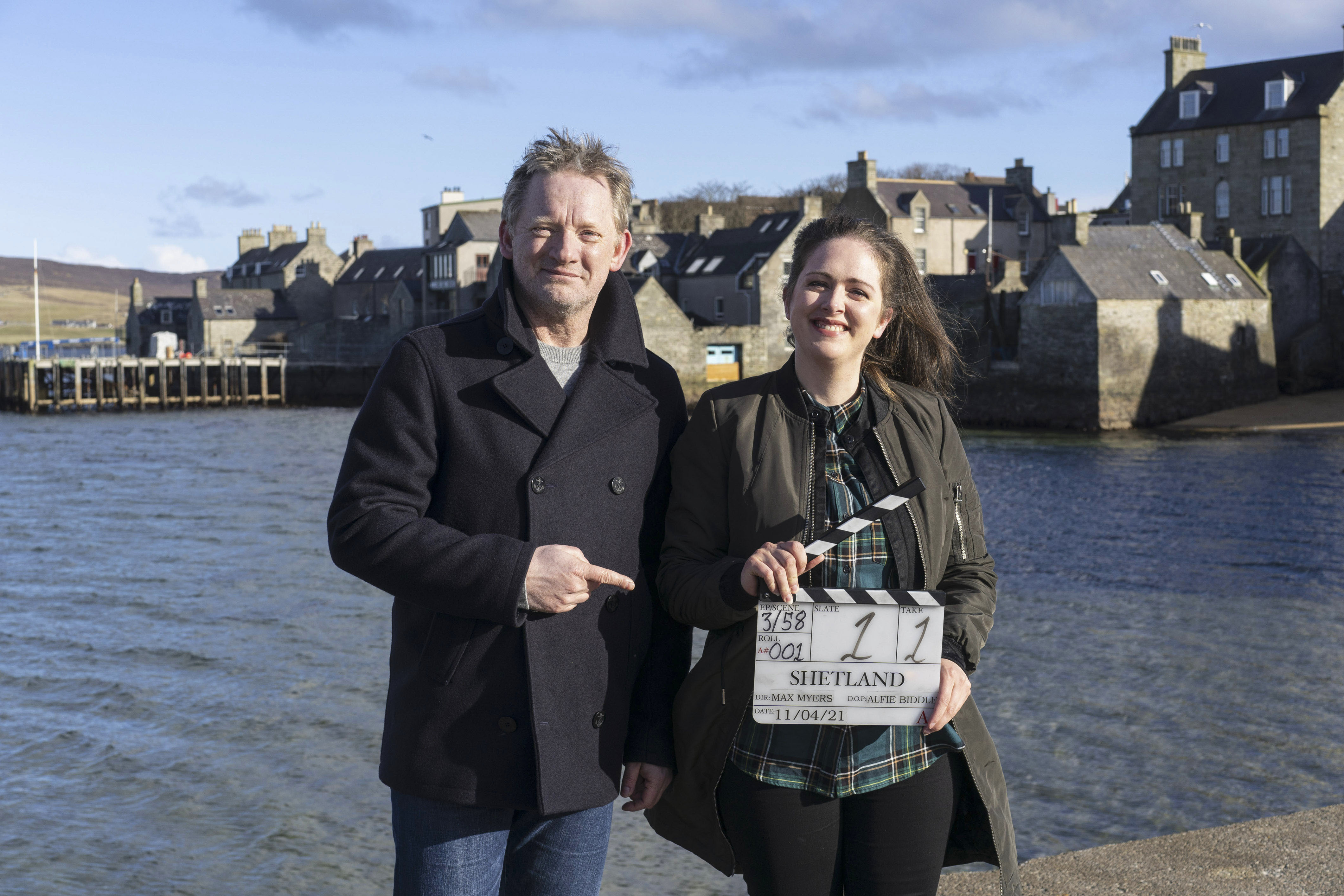A man and a woman smile standing at the sun with a clapboard that says 'Shetland'. A body of water and buildings can be seen behind them.