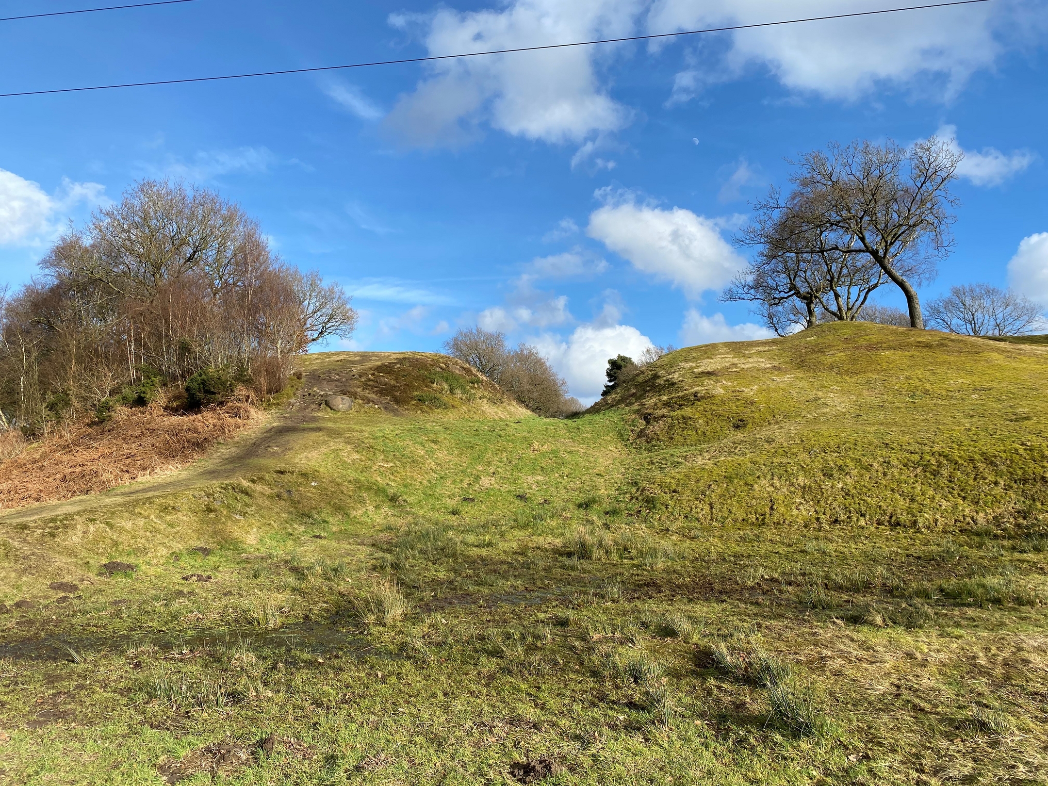 A ground-level view of grassy earthworks at Rough Castle, showing a sunlit dip between two rounded mounds. Trees and shrubs line the edges under a bright blue sky with scattered clouds.