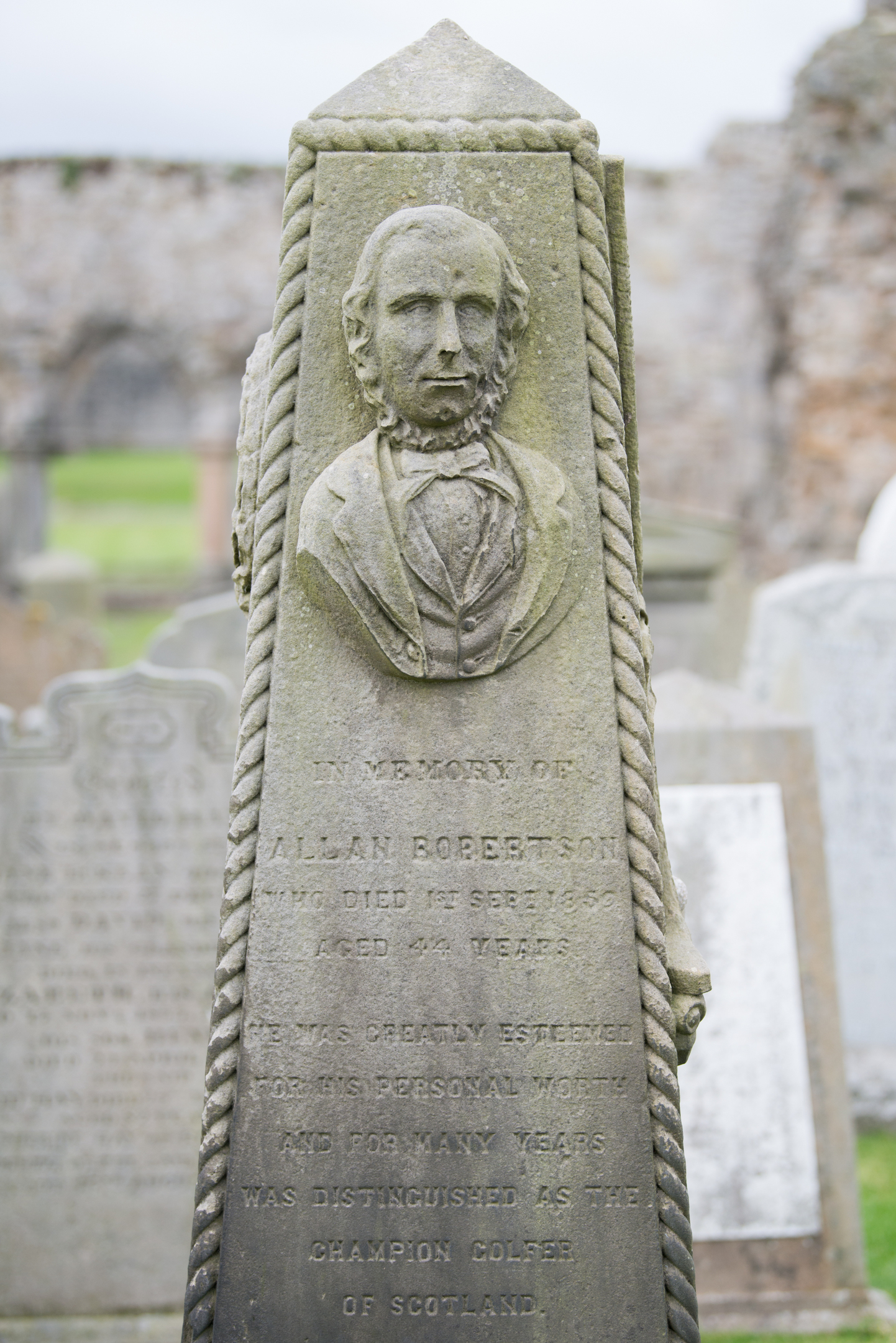 A small obelisk marking the grave of Allan Robertson. It features a striking and intricate stone carving of Robertson.