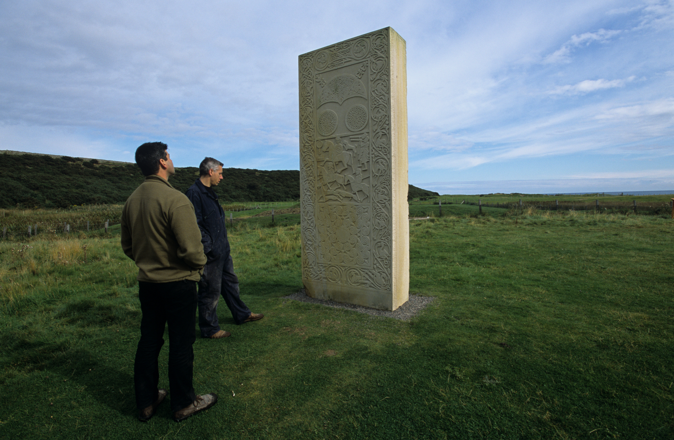 Two visitors standing in front of the Hilton of Cadboll replica stone, observing its carvings. The stone is positioned on a grassy area with hills in the background.