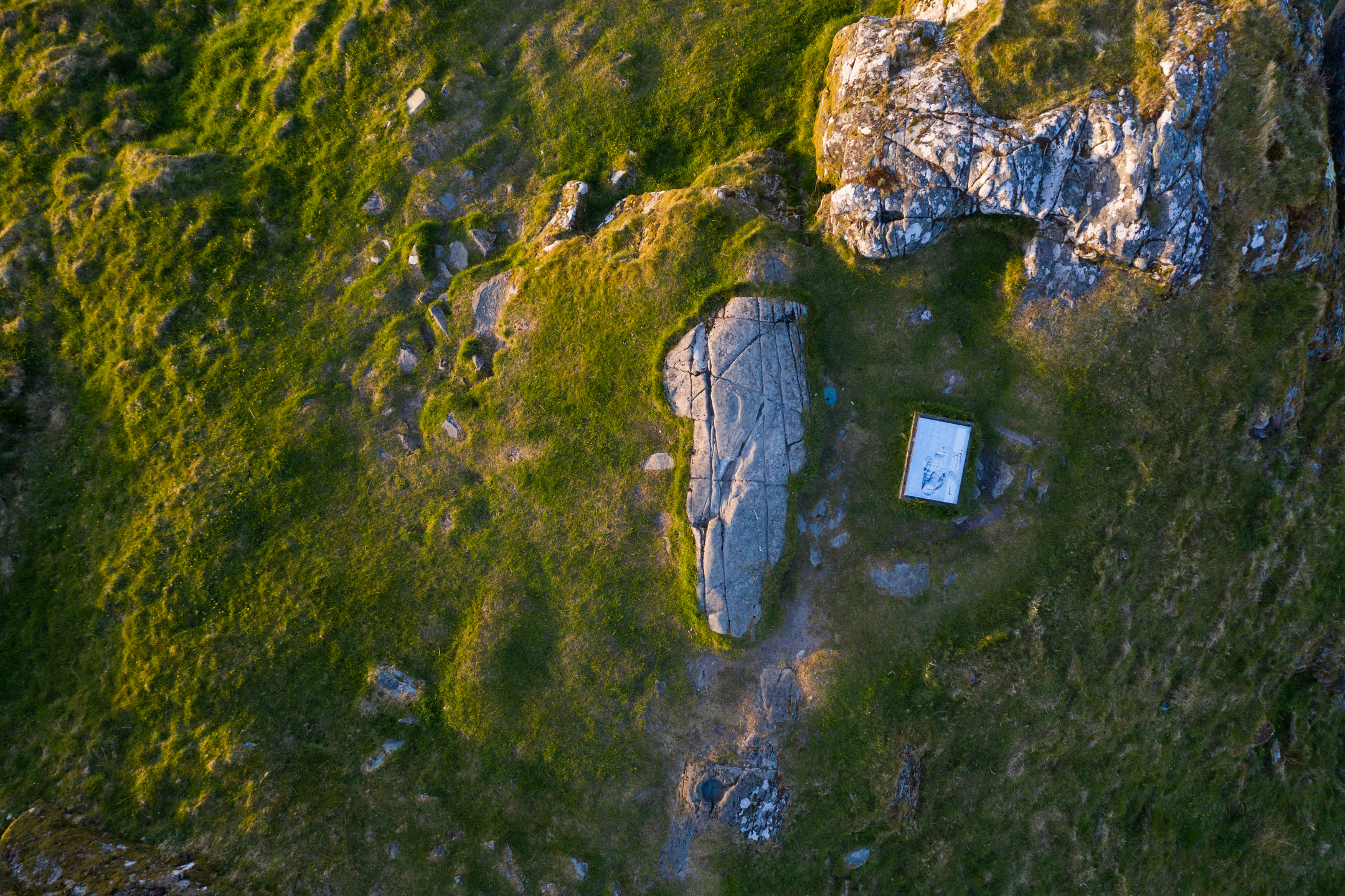 Aerial view of the carved footprint stone on Dunadd Hill, surrounded by rough rocky and grassy ground. There is an information panel beside the rock.
