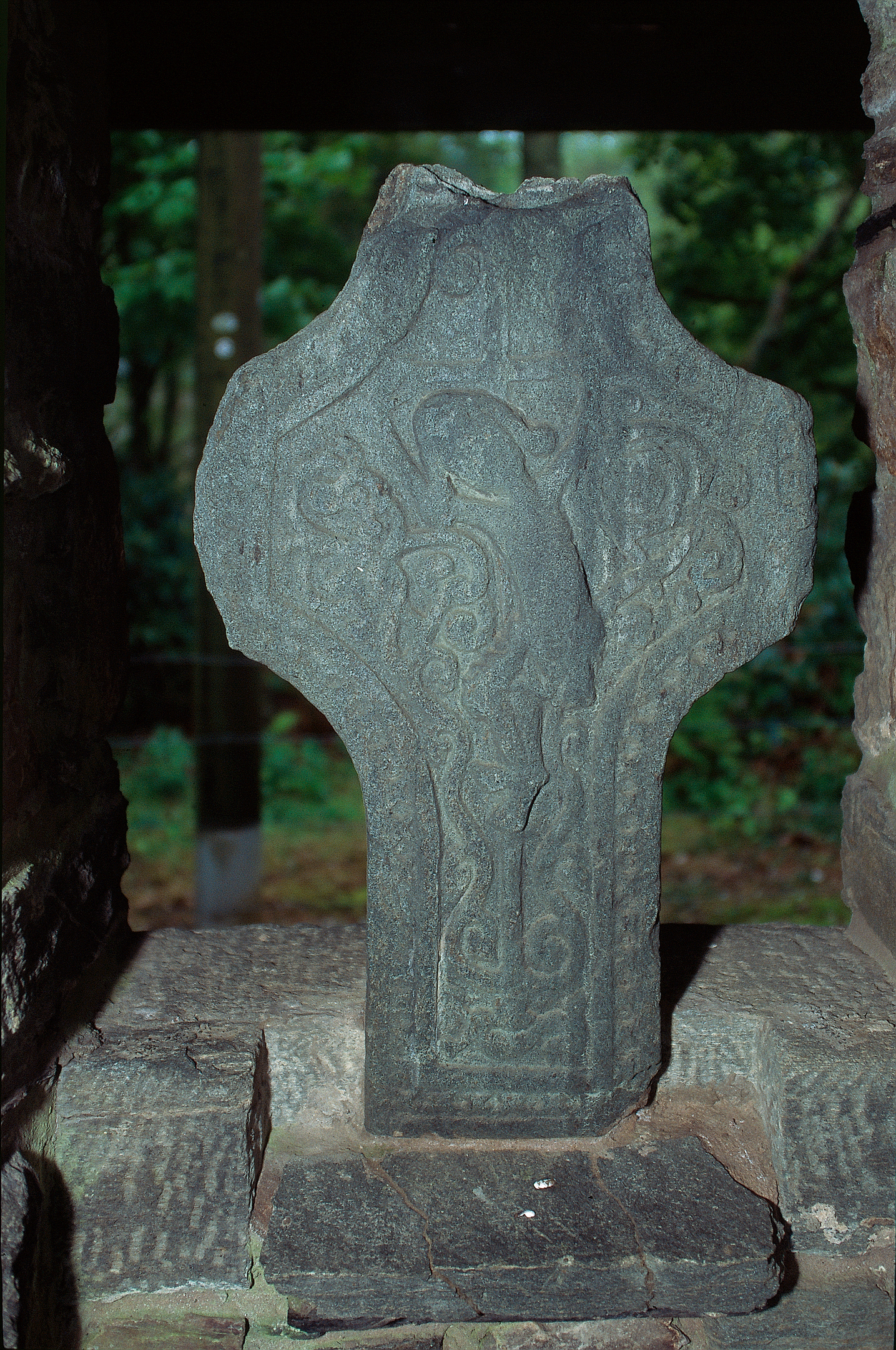 Carved stone on a plinth featuring a figure with greenery in the background. 