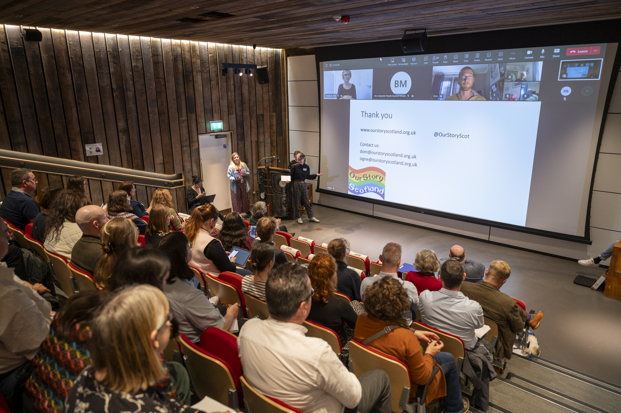 Attendees at a conference-style event sit in rows of banked seating while a presenter delivers a speech in front of a big screen,