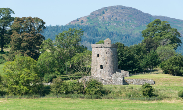A rounded stone tower surrounded by low ruinous stone walls stands in a green field. There are green leafy trees surrounding the building and a mountain stands in the background behind.