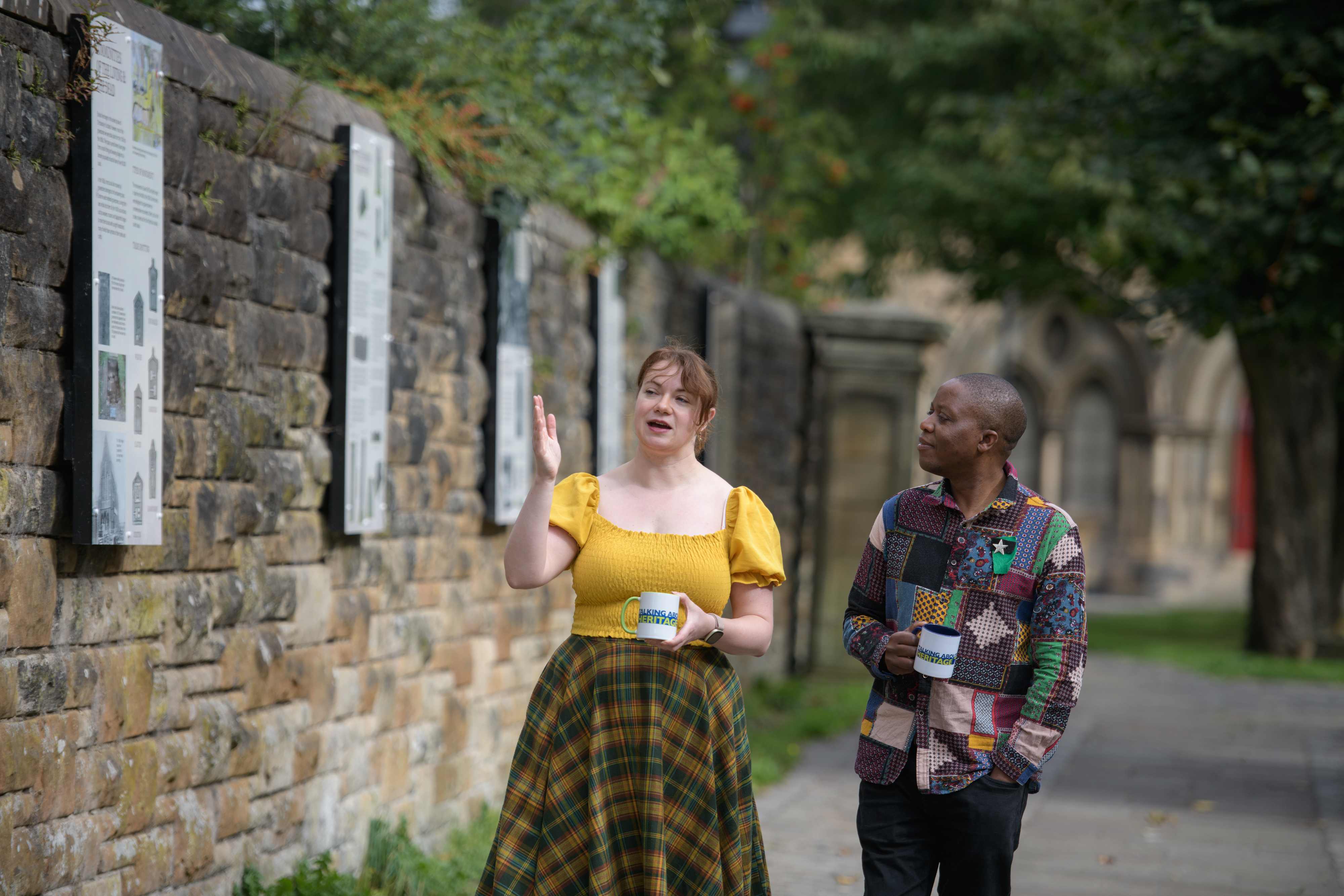 Two people walk alongside an old stone wall talking and holding white mugs with the Talking about Heritage logo on them. In the background out of focus, there is what appears to be an old church.