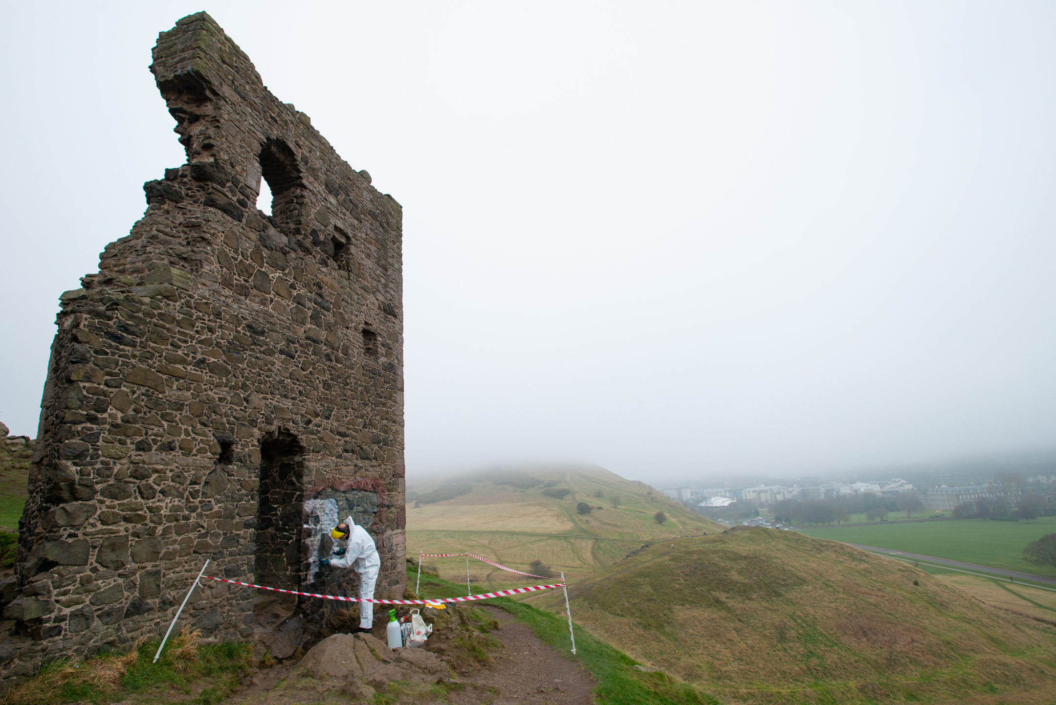 A worker in white overalls works on removing graffiti from a historic chapel wall, on a bleak, wet day. 