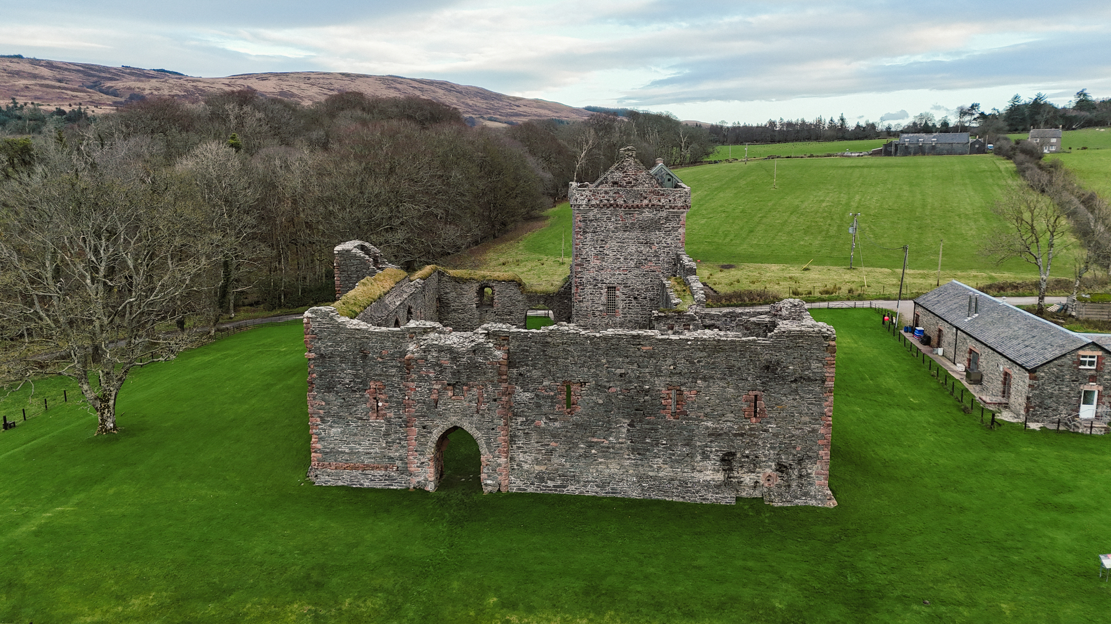 Drone view showing the front and side walls of a medieval castle ruin, bordered by green fields and woodland.