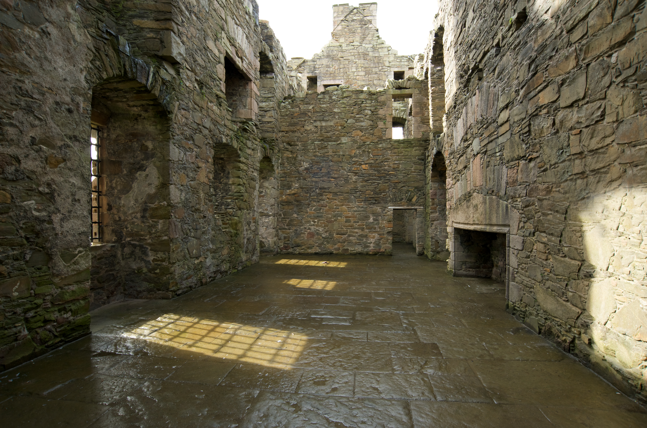 An interior room of a ruined stone castle, with bare stone walls, empty window openings, and sunlight casting grid patterns on the worn stone floor.