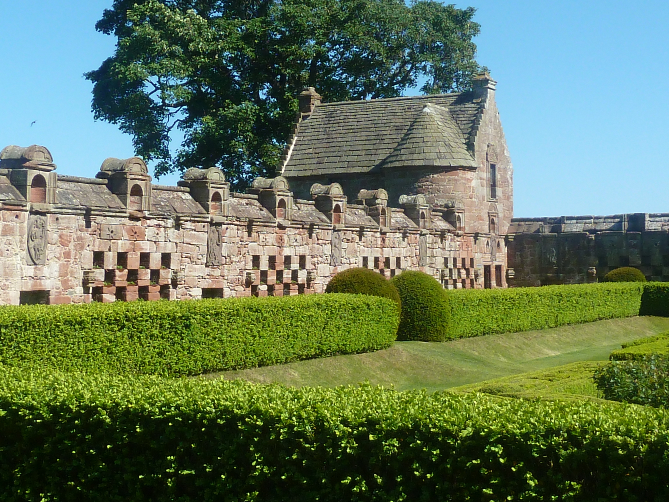 A well-maintained walled garden with neatly trimmed hedges. The garden is enclosed by a distinctive stone wall with small turrets, carved panels and recesses perhaps once used for storing beehives.