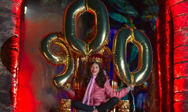 A young girl with long brown hair sits atop a throne wearing a crown. She smiles at the camera, wearing a pink coat, pink scarf and holds large gold balloons that spell out the number 900. Behind her is a stone archway lit up with red lighting and fog.