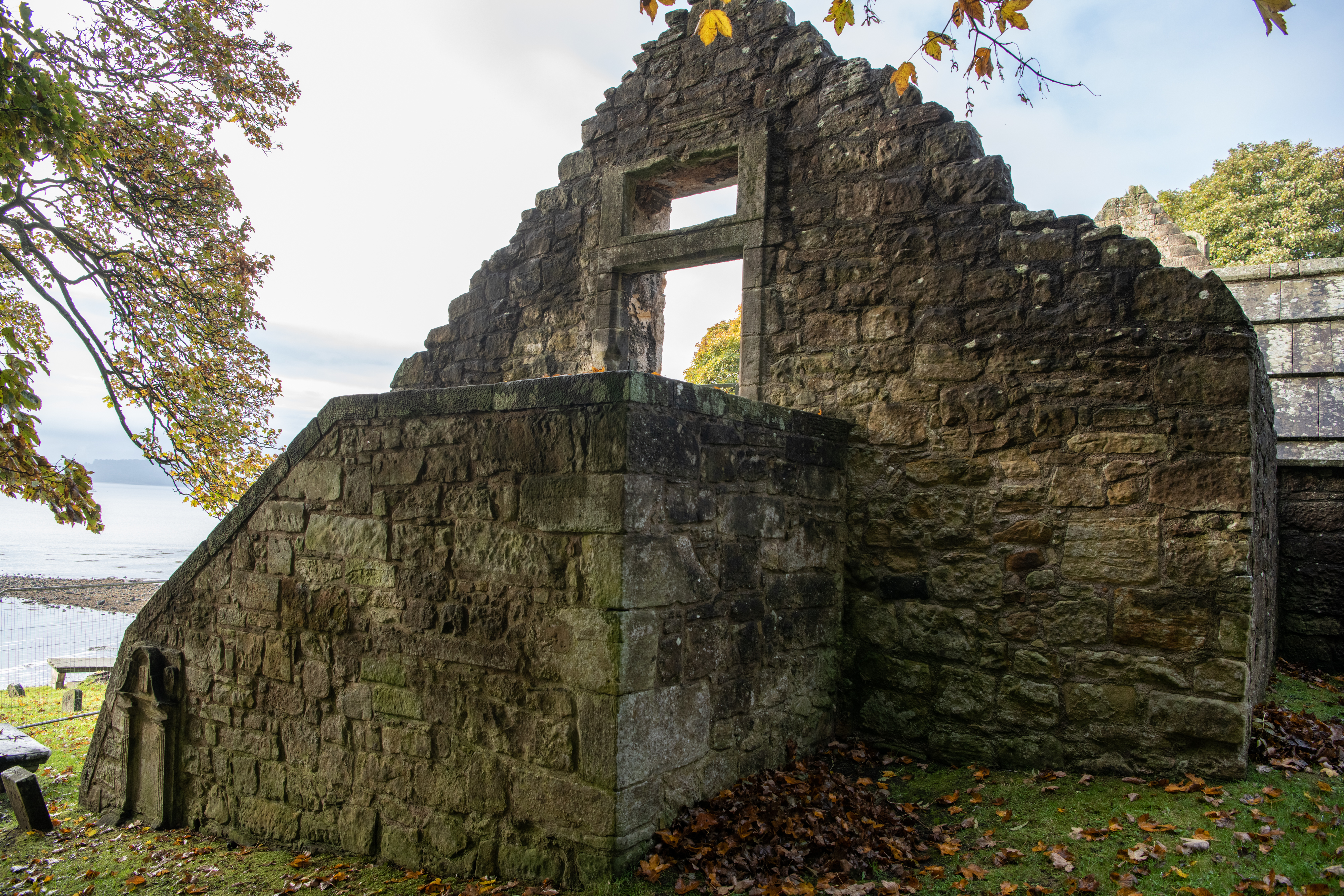 A close view of the church ruins showing weathered stone walls, arched doorways and window openings, with autumn leaves scattered across the grass.