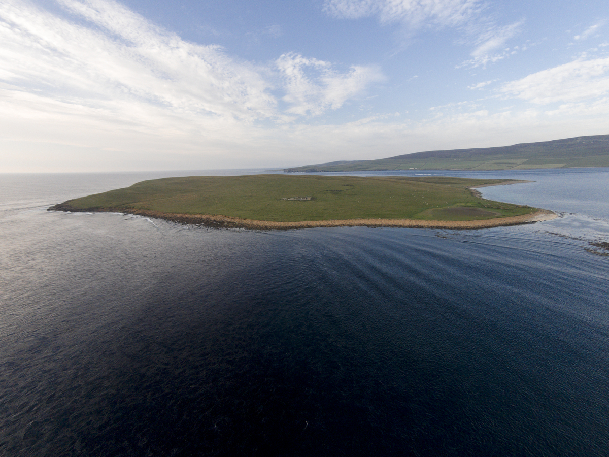 Aerial view of open, grassy landscape of a tiny island bordered by sea, with the faint shape of a ruined building just about visible