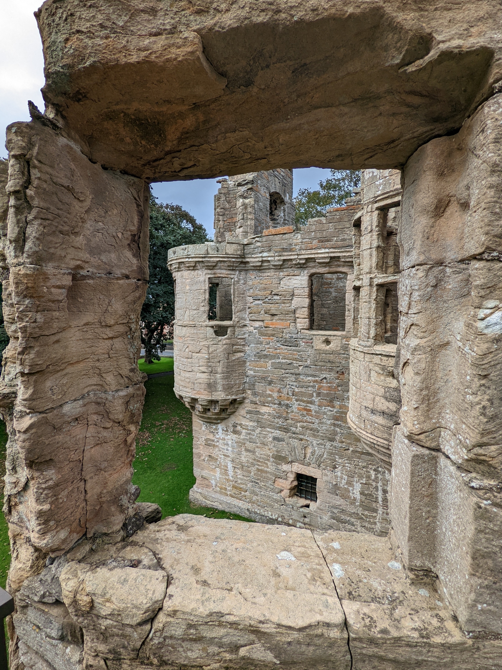 View of a stone tower and turrets framed through a rough stone window opening, with green grass and trees visible below.