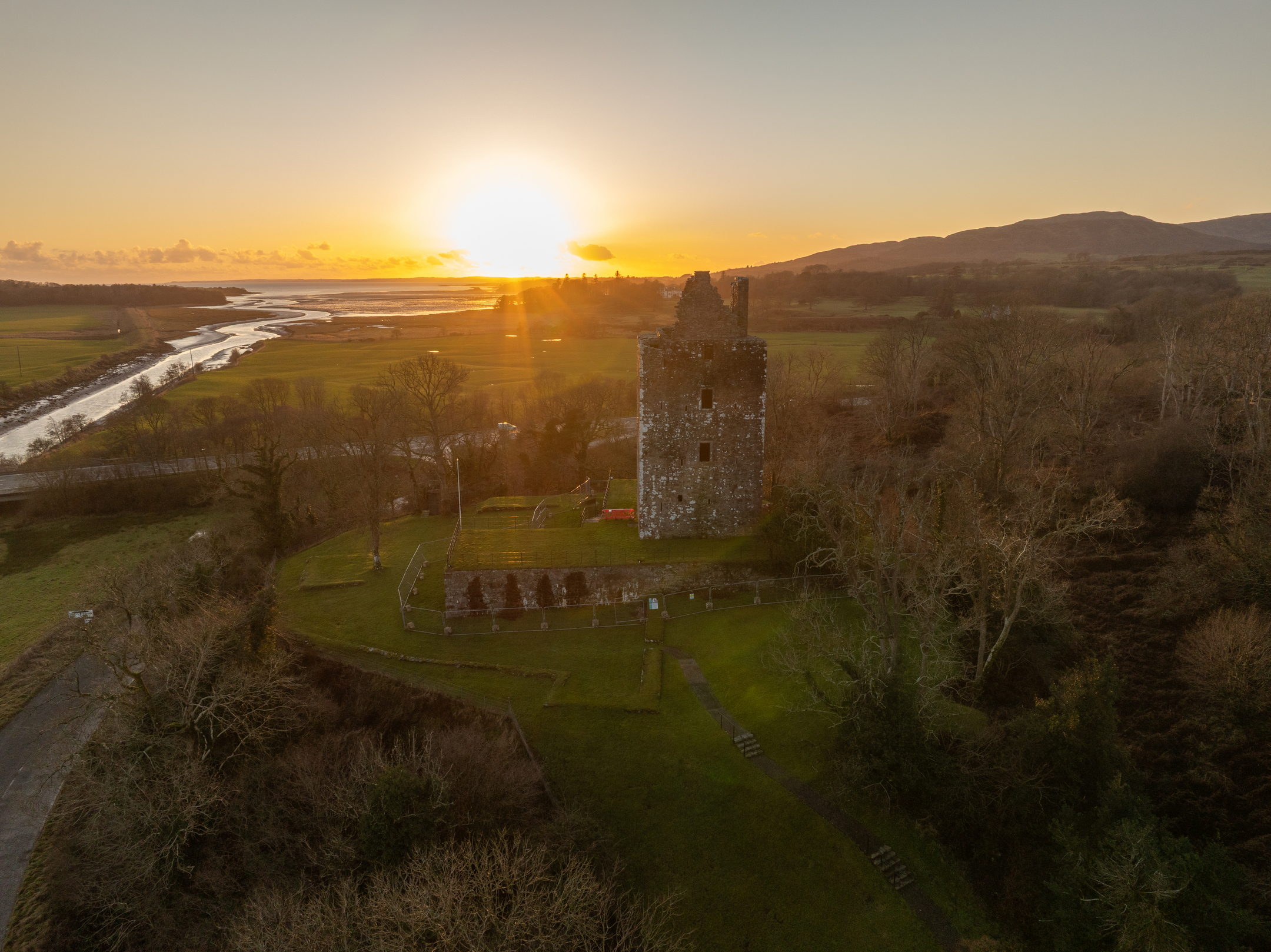 A drone‑view of a ruined stone tower standing on a grassy hill at sunset. The sun is low on the horizon, casting a golden glow over surrounding fields, a winding river, and distant hills.