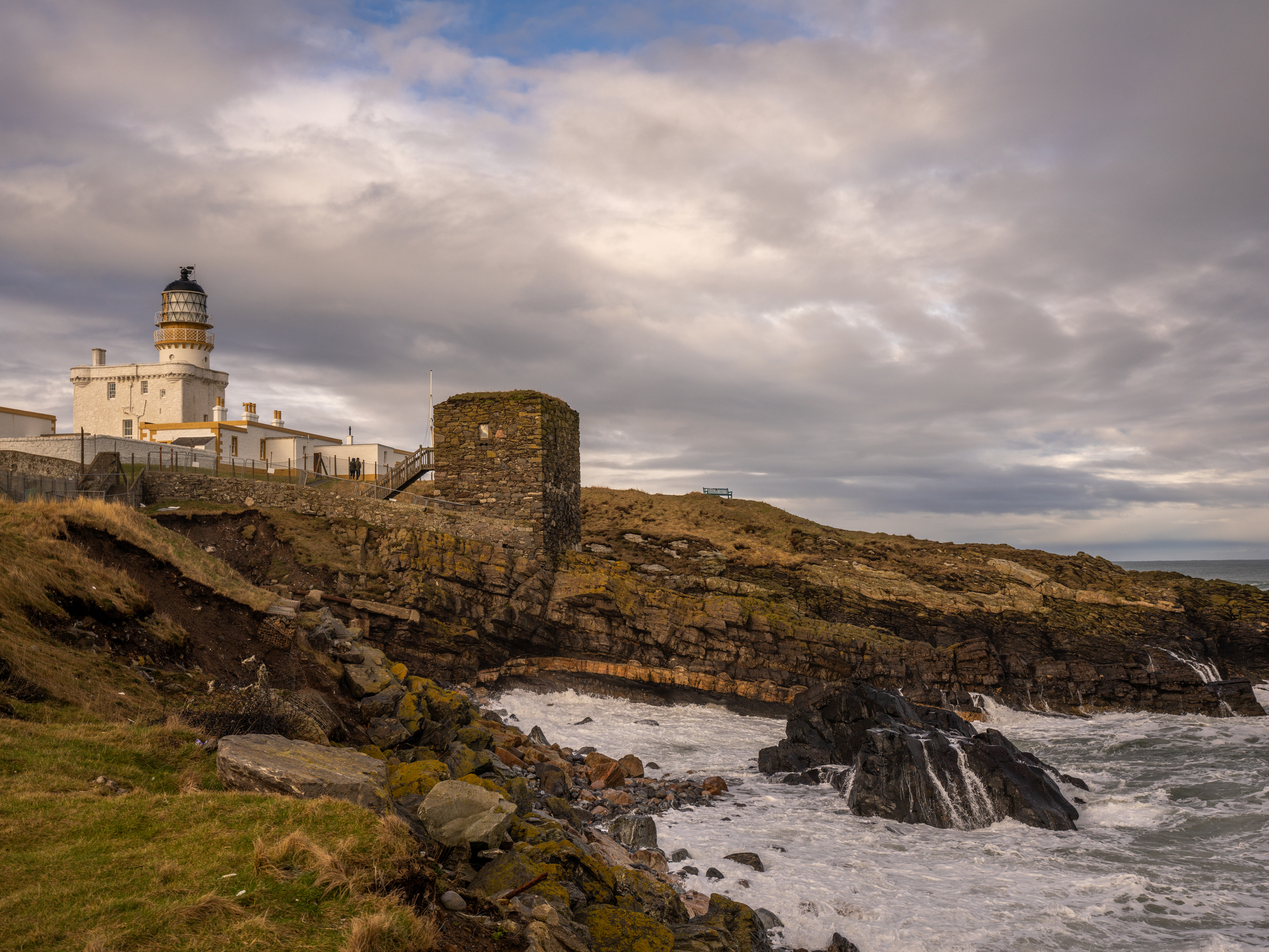Clouds gathering above a historic lighthouse. In the foreground there is a rocky shoreline and a small, square stone building.