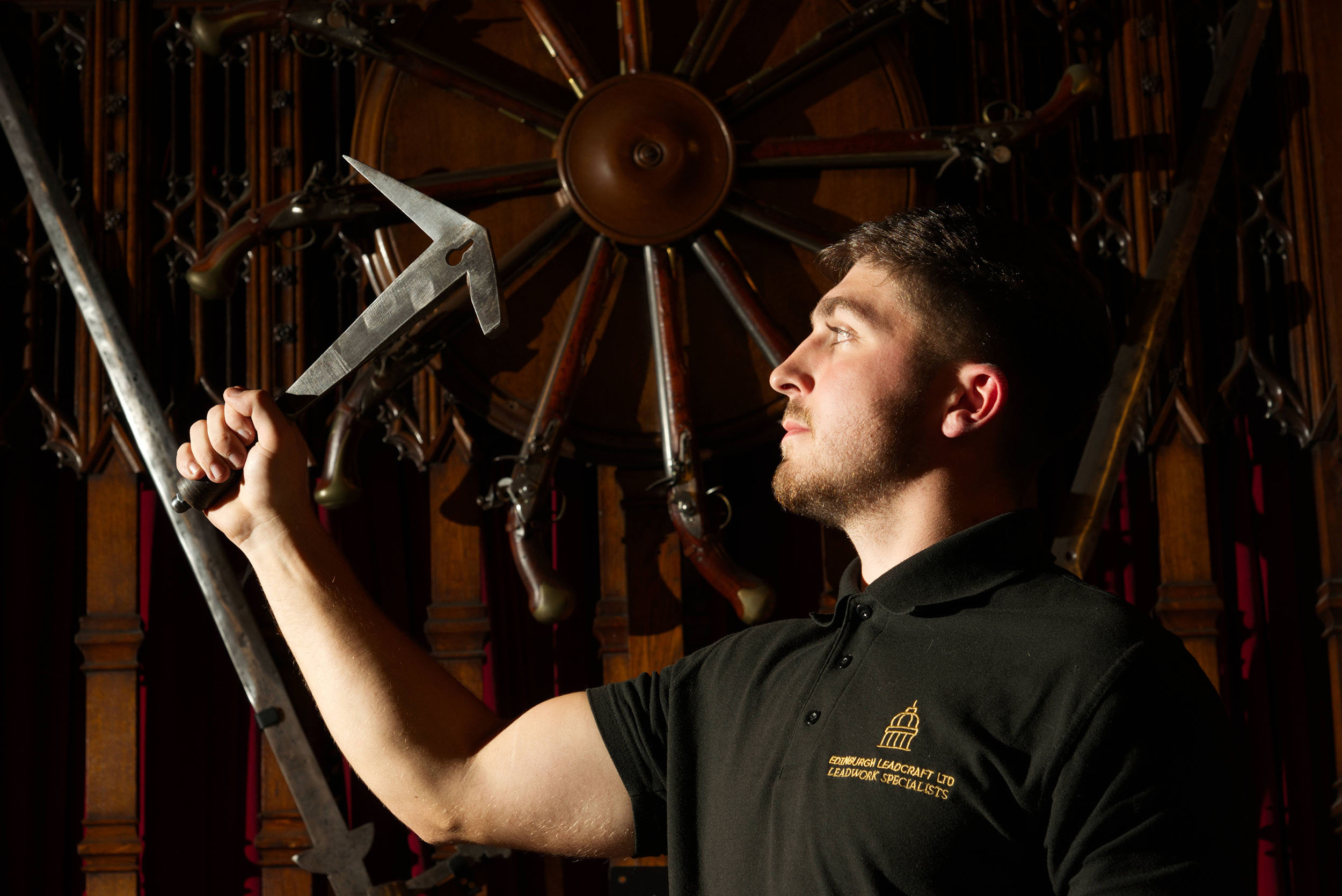 Man holding a leadworking tool, wearing a black branded polo shirt, lit from the side against a dark background