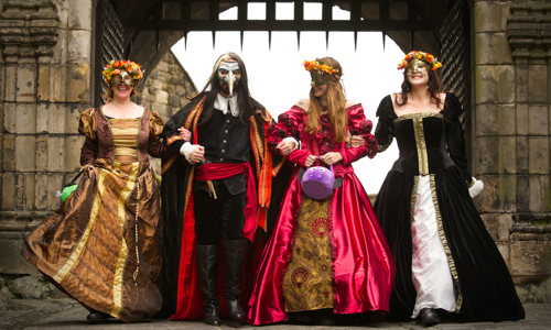Four costumed performers walk towards the camera out of the gate of Edinburgh Castle, linking arms and smiling. Three wear historical dresses in gold, red and black, and one (second from left) wears black trousers with a black shirt, cape and red sash