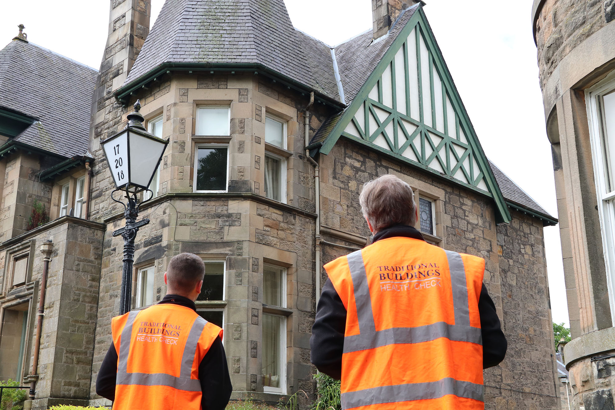 Two people wearing high-visibility orange vests with "Traditional Buildings Health Check" printed on the back stand facing a large stone building. The historic building features bay windows, steep slate roofs, chimneys, and a green-and-white timber gable. A traditional street lamp marked "1720 18" stands in the foreground.
